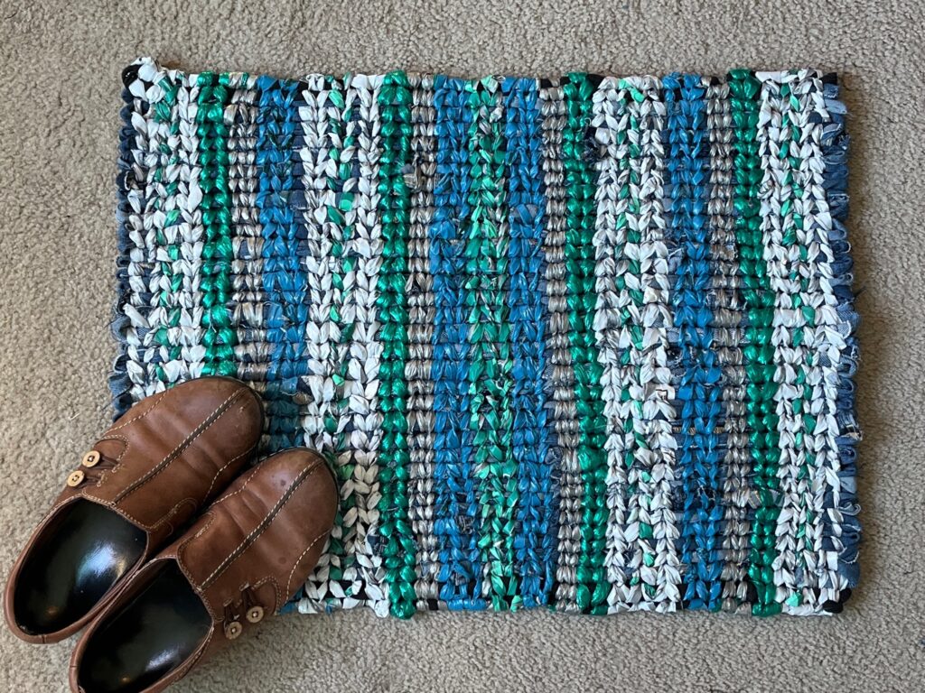 Finished rug in white, green, grey, blue and dark green with a pair of brown shoes on it.  